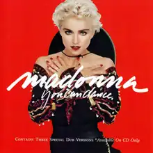 Madonna with short, blond hair, wearing a black dress, with her hands folded against her breasts and standing in front of a red background. She has a Spanish hat attached to her neck.